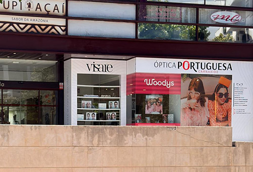 Storefronts with signage: Óptica Portuguesa and Woody's, glass display windows with eyewear images, beige concrete barrier in front.