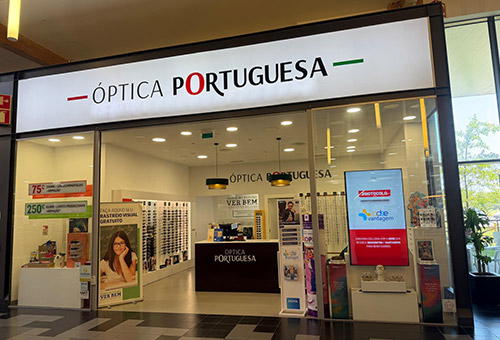 Front of Óptica Portuguesa eyewear store with bright white signage and green/red accents; interior displays wall of glasses and a central checkout desk.